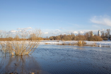 March sunny day by the river. A picturesque landscape, early spring, a river with snow-covered banks, dry grass and bushes. Church in the background. The first thaws, the snow is melting