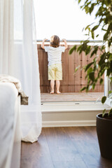 A little boy stands on a balcony at home or in a hotel room and looks into the distance. Expectation. Leisure and travel.