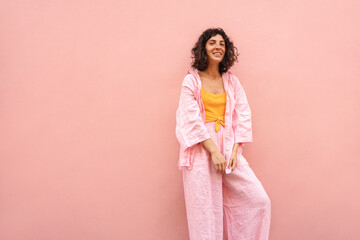 Obraz premium Beautiful young caucasian woman is smiling looking into camera on pink background with place for text. Brunette with bob haircut wears summer shirt and pants. Happy weekend concept. 
