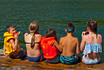 Children eat watermelon on the pier. Children in life jackets rest on the river bank. Camping by the water.