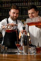 bartenders preparing cocktails, one is pouring drink into jigger and other pouring cocktail into glass