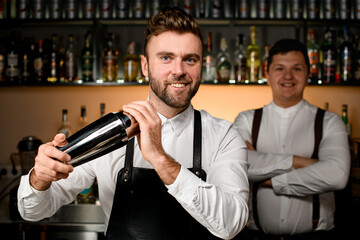 Young smiling bartender shaking a cocktail in steel shaker in bar interior