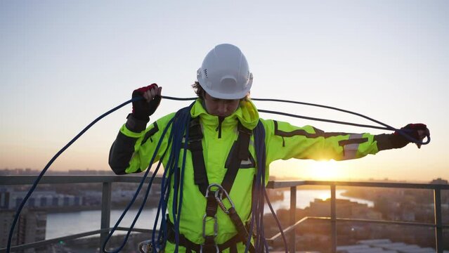 Industrial climber on rooftop untangles safety rope and hangs skein on shoulders. Man in light green overall and helmet prepares for work at sunset