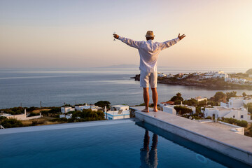 A happy man with a drink stands by the swimming pool and enjoys the view over the sea during a...