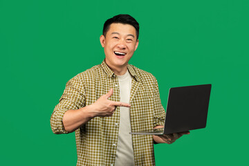 Happy asian middle aged man pointing at laptop computer and smiling at camera on green studio background
