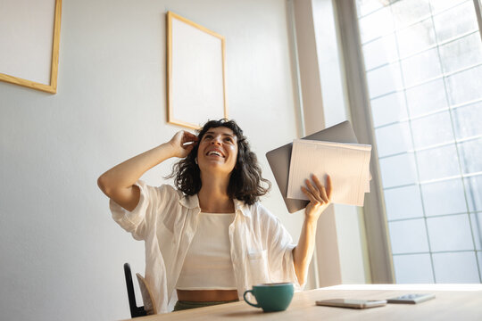Joyful young caucasian woman looks up holding work papers sitting at her desk. Brunette wears white shirt on casual day. Concept of positive emotions 
