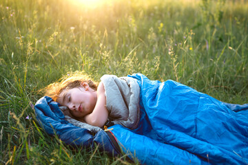 A child sleeps in a sleeping bag on the grass in a camping trip - eco-friendly outdoor recreation,...