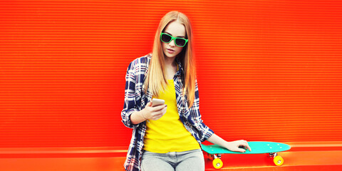 Modern blonde young woman with smartphone and skateboard on orange background, blank copy space for advertising text