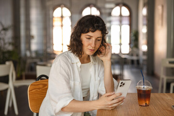 Pretty young caucasian girl reads news on Internet on her phone sitting at her desk. Brunette wears casual summer clothes sitting in cafe. Relaxed lifestyle, concept