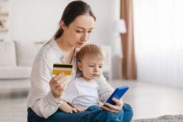 Pretty young mother and little kid son buying something on Internet, using cellphone and holding credit card