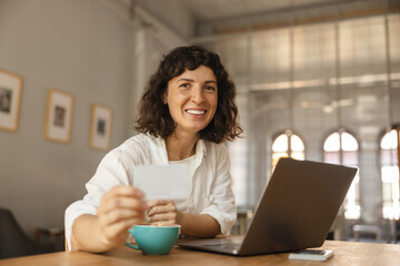 Pretty young caucasian woman makes online money transfer sitting at laptop in cafe. Brunette woman in casual clothes smiles as she looks at camera. Lifestyle concept