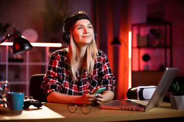 Charming woman using smartphone and headphones for enjoying favorite song. Young lady in cap taking break while working on laptop at home. Freelance and technology concept.