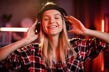 Naklejka premium Pretty woman and listening music in holding headphones in hands. Young lady in checkered shirt sitting at desk with wireless laptop. Relaxation during evening work at home.