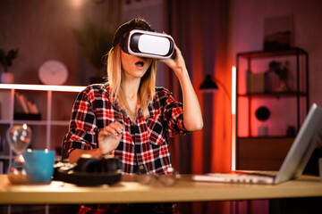 Young attractiv woman sitting at home office and using VR headset and laptop for work during evening time. Concept of people, technologies and freelance.