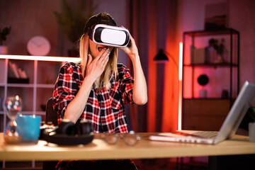 Young attractiv woman sitting at home office and using VR headset and laptop for work during evening time. Concept of people, technologies and freelance.