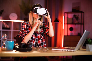 Young attractiv woman sitting at home office and using VR headset and laptop for work during evening time. Concept of people, technologies and freelance.