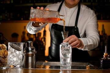 Close-up view of male bartender hand holding crystal bottle and pours beverage into steel jigger