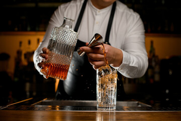 close-up of glass with ice cubes into which bartender pours drink from steel jigger