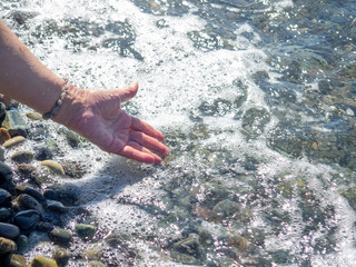 A hand with a bracelet made of stones reaches for the water. Sea wave and female hand. Feel the wave. Pebble beach.
