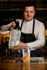 male bartender holds a siphon and splashes on glass with a cold cocktail