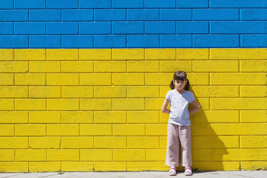 little toddler girl sad on the background of a brick wall painted in the colors of the Ukrainian flag, the concept of supporting the children of Ukraine during the war, place for text