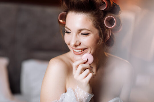 Beauty portrait of a beautiful smiling young brunette woman with bright pin-up make-up eating delicious french macaroons or macarons. girl in white dress and pink curlers sitting on the bed and relax