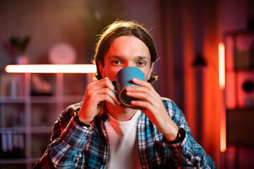Confident young man drinking coffee or tea or hot drink at office. Caucasian male worker working at modern office or home at the evening time.