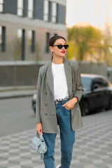 Trendy girl in sunglasses wearing grey jacket and jeans walking in city. Pretty caucasian woman spend time outdoor 