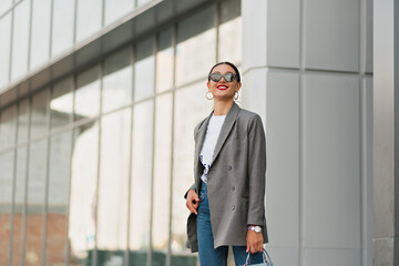 Brunette european woman in grey jacket and jeans in sunglasses walks outdoors. Stylish lady smiles sincerely and moves outside.