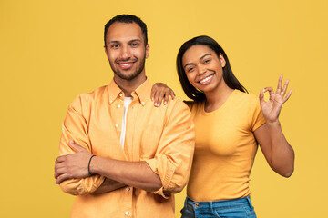 Happy young black couple showing okay gesture, approving or recommending something over yellow studio background