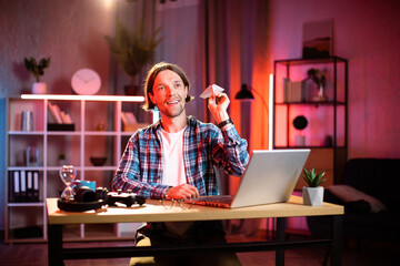 Happy young man in casual wear throwing paper plane while taking break during remote work on laptop. Smiling caucasian male sitting at desk during evening time and having fun.