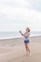 Mature woman with blonde hair wearing sunglasses taking a photo with smartphone on top of a dune. Beach and sea behind.