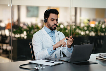 Confident busy young muslim male therapist with beard in glasses, headphones and white coat speaks with patient