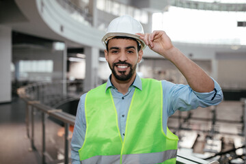 Happy millennial middle eastern builder engineer with beard in protective clothing touches hard hat...