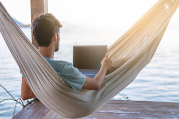 young digital nomad lying on hammock with sea views working remotely on internet at sunset - Traveling with a computer - Dream online job concept - Selective focus.