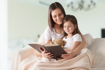 Happy mom and teen daughter smiling and reading interesting book while relaxing on weekend day at home