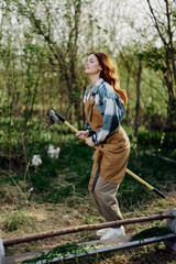 The woman hops amusingly with her rake like a witch on a broomstick in her green garden among the trees and smiles. The concept of everyday lifestyle and joy in the moment