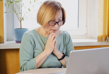 Socked surprised mature woman in eyeglasses looking at laptop reading disturbing news in media, watching video content, sitting indoors. Feeling hope, praying for happy end. Received bad email