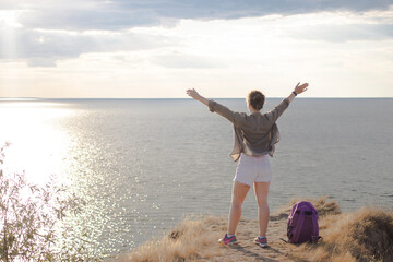 Back view of happy woman at mountain peak with open raising arms celebrating, hiker standing on rock during sunset and looking at sea. Strong and freedom lifestyle concept. Enjoying adventure travel