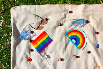 LGBTQ picnic in nature in summer park. White plaid on green grass, pride month symbols - LGBT flag, dishes, blue glasses, cherries contrast shades view from above. Colorful objects. Love and freedom.