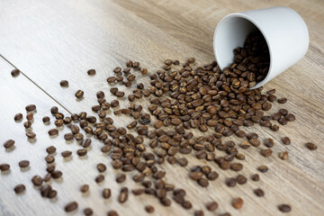 A cup filled with roasted coffee beans, which fell on the wooden table, scattered the coffee beans on the table.
