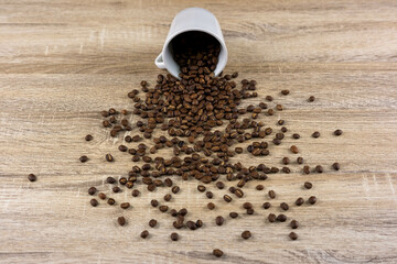 A cup filled with roasted coffee beans, which fell on the wooden table, scattered the coffee beans on the table.