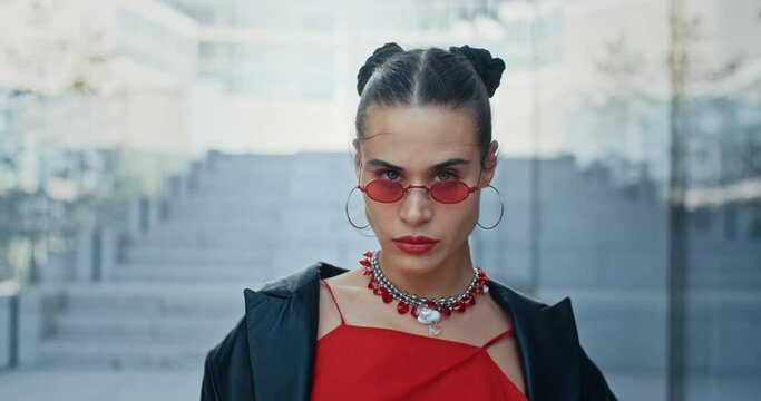 Slow motion portrait of stylish serious woman, seductive urban style female in cool red eye glasses looking at camera standing in wind in modern business center overlooking high-rise glass buildings