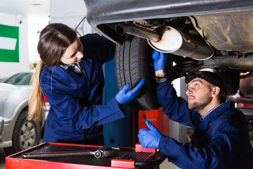 positive female and master are standing near car and repairing it in workshop.