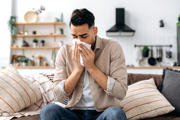 Unhappy sad indian or arabian guy suffering from fever and flu, sitting on sofa in living room,...