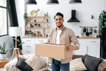 Joyful stylish indian or arabian guy, stand at home in the living room, holding a large cardboard...