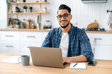 Portrait of a handsome successful confident Arabian or Indian guy with glasses, freelancer, designer or IT specialist, sitting at home in the kitchen, working online by laptop, looks at camera, smiles
