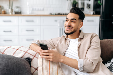 Positive handsome indian or arabian guy in trendy clothes, sitting in a modern living room on comfortable sofa, holding his smartphone, texting message, browsing social media, surfing web, smiling