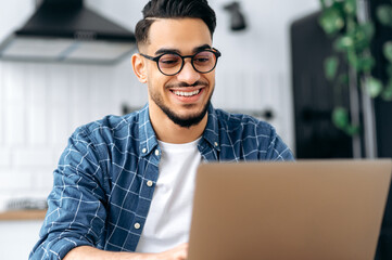 Close-up photo of attractive positive indian or arabian guy with glasses, freelancer or creative designer, sitting at a desk at home, working on a creative project uses laptop, looks at screen, smiles
