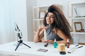 Cheerful tanned handsome curly Latin beauty blogger in casual top present favorite spray in home interior. Copy space Mockup Banner. Influencer record video review blog using smartphone